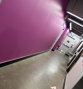 Washrooms Hallway in the J-Wing. Has mens womens and accesibility washroom. Also has some classes in this hallway as well as a water fountain. One wall is painted Purple. Lights are hanging above, semi narrow hallway.